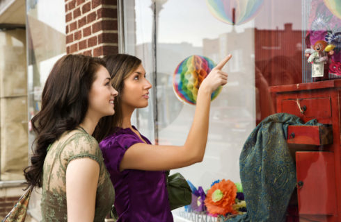 Two young brunette women in short-sleeve bloulses look at a antiques shop window.