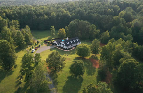 Arial view of large farmhouse with a black roof and dormers, with a swimming pool and manicured lawn.