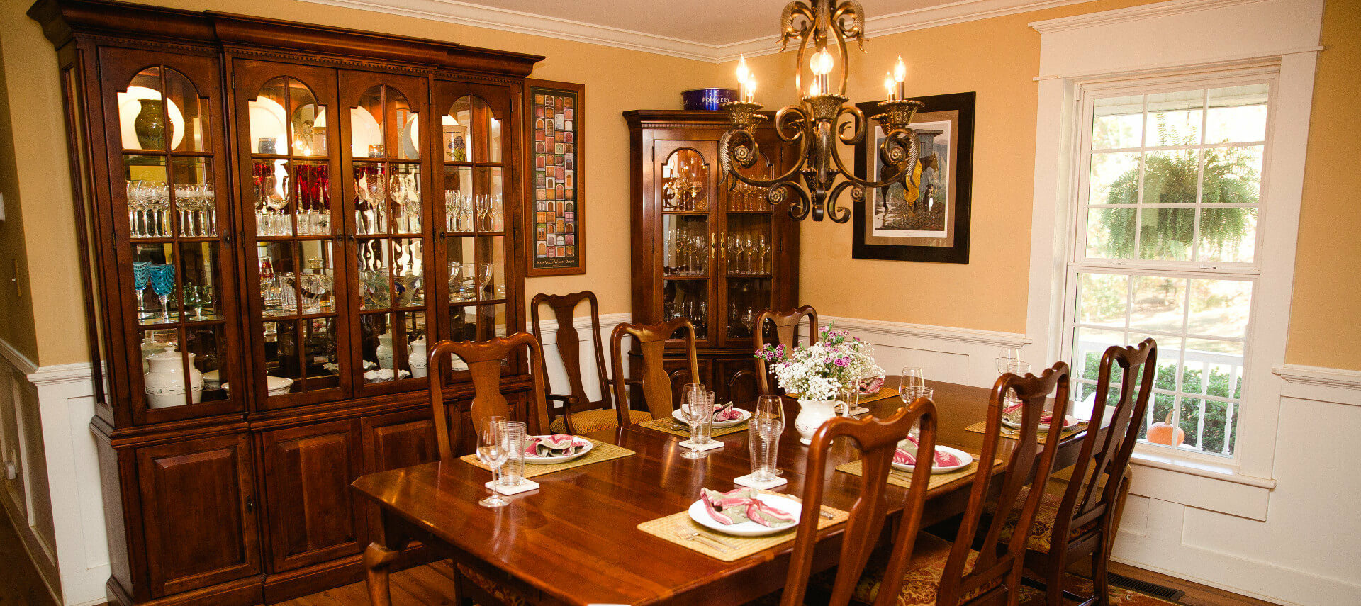 Dining table set for six in a warm and bright dining room with glass china cabinet and corner hutch by window with and wooden floors.