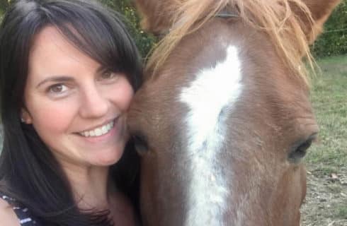 Pretty brunette lady snuggles close to a brown horse with a white blaze in a selfie.