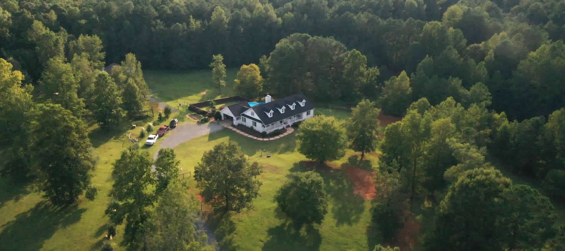 Arial view of large farmhouse with a black roof and dormers, with a swimming pool and manicured lawn.