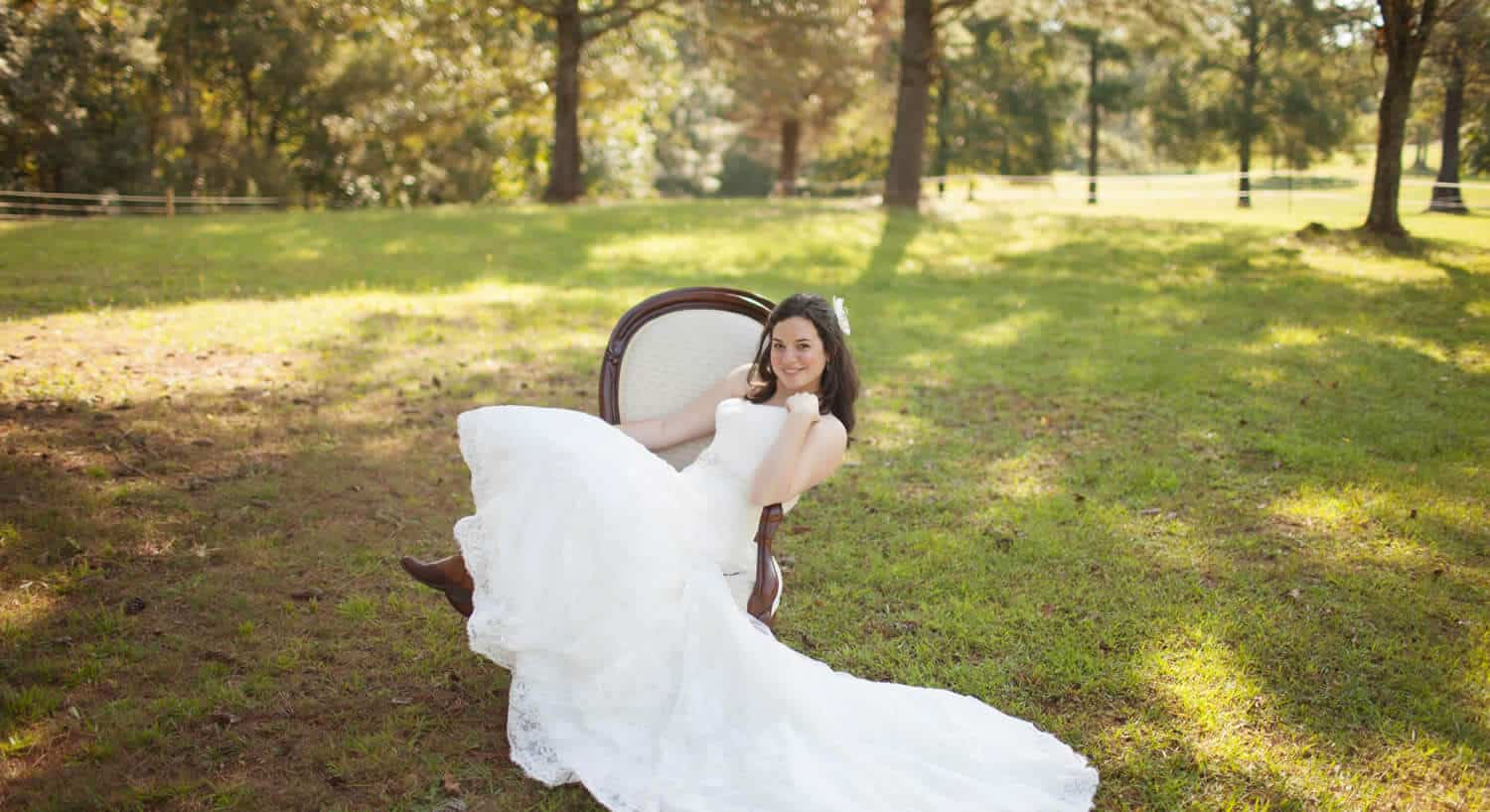 Bride in a white lace gown and brown cowboy boots leans back in a chair set in a pasture. 