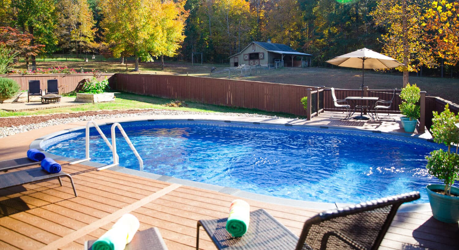 In-ground pool surrounded by wood deck with lawn chairs facing cream barn with green roof and trees all around