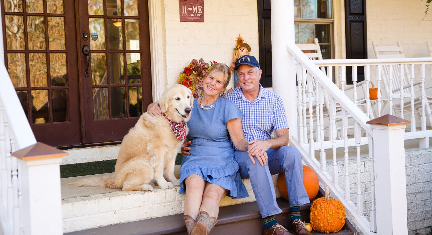 Man and woman wearing blue attire sitting on steps of inn with fall decorations and golden retriever beside.