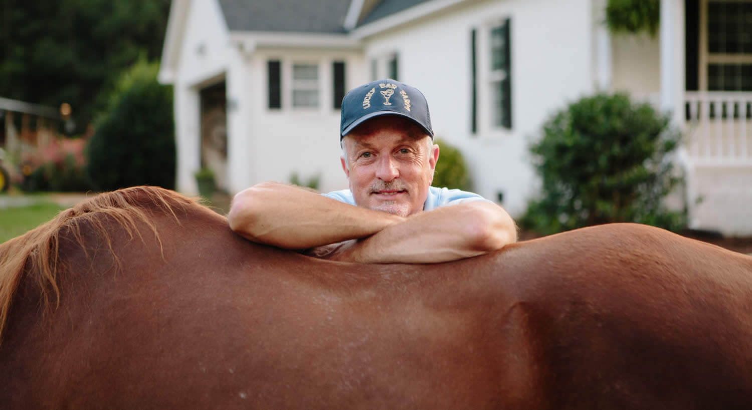 Man in a blue ball cap leans on the back of a brown horse. 
