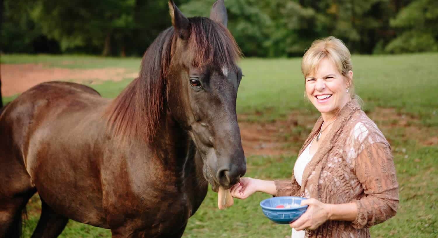 Smiling woman is feeding treats from a blue bowl to a brown horse.