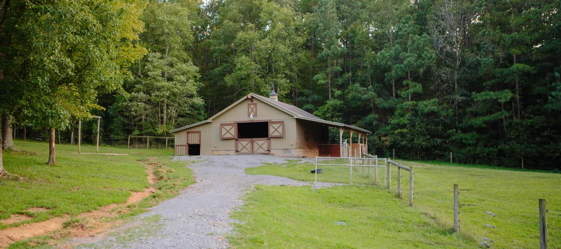 Brown horse barn with tan accents at the end of a gravel drive, set in a green pasture.